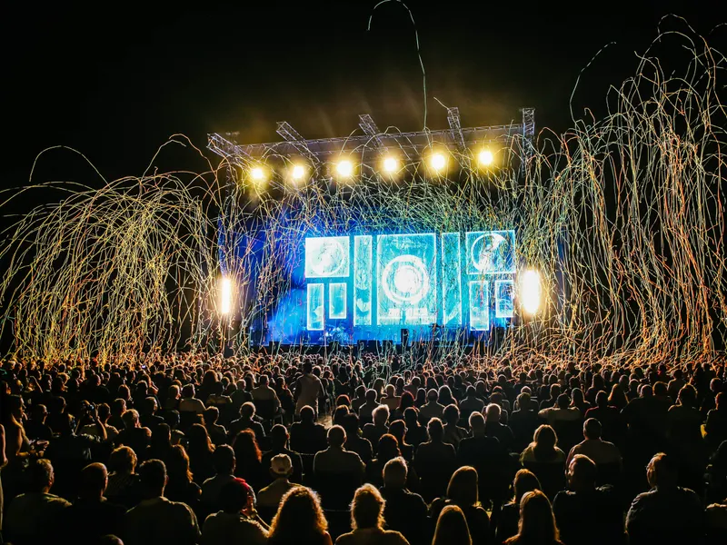 A packed open-air crowd facing a lit stage as gold sparks fall from the rig.