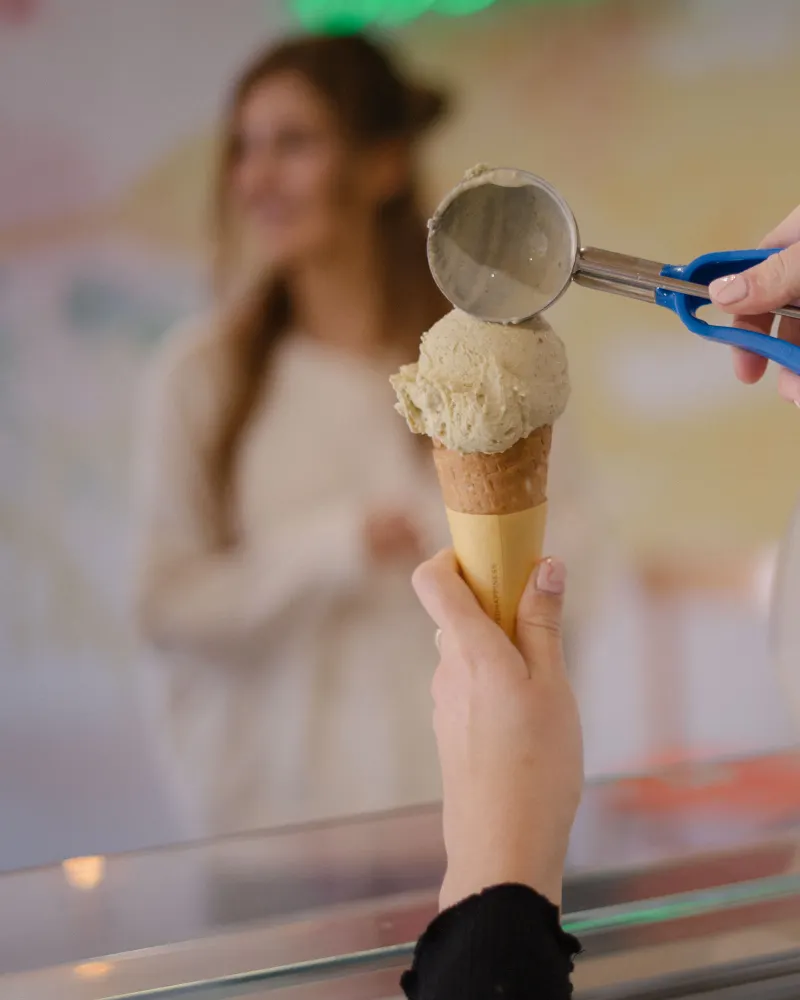A server presses a scoop of pale ice cream onto a sugar cone across the counter, a customer waiting behind.