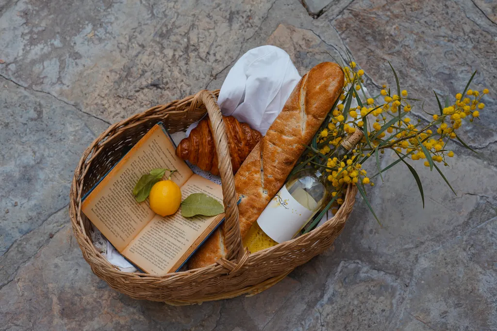 A wicker basket with a baguette, lemons, a book and a sprig of yellow mimosa.