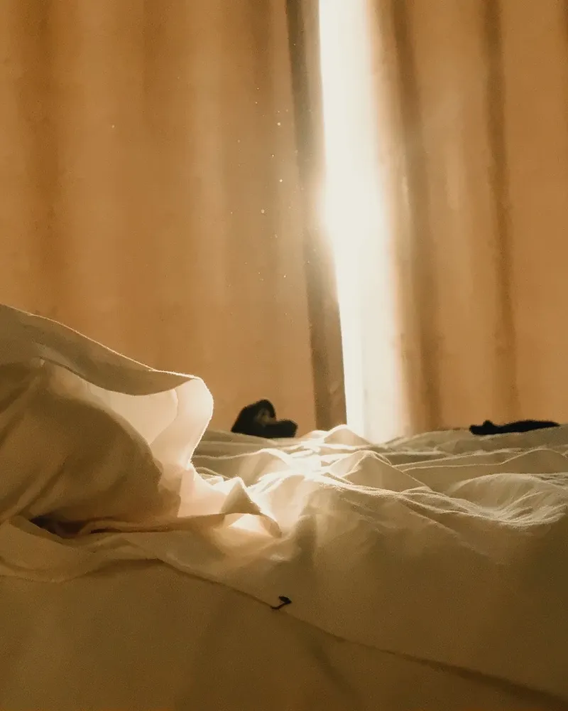 Morning light through linen curtains onto an unmade white bed.