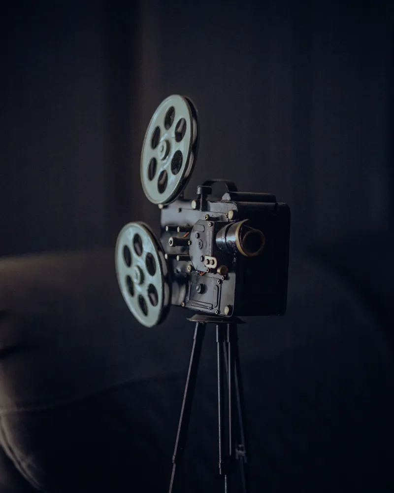 A vintage film projector with two reels on a tripod, against a dark curtain.