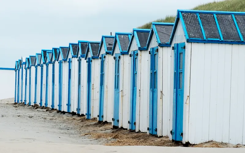 A long row of white beach huts with blue doors on the sand.