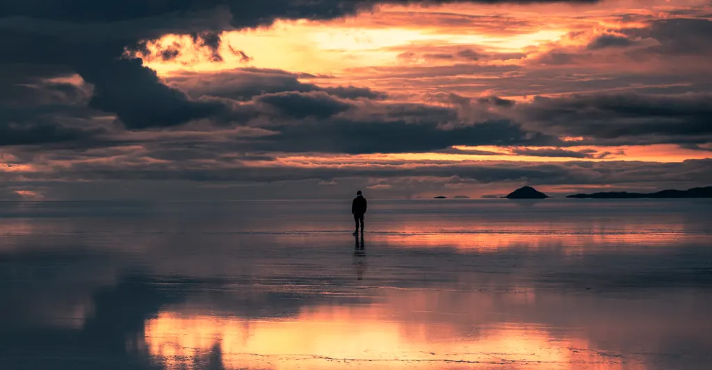 A lone figure stands on wet salt flats that mirror a low red sunset.